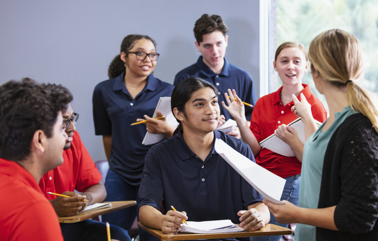 A multiracial group of six high school students sitting together in a classroom having a discussion, with a teacher leading. The students are in two teams, wearing either blue or red shirts. They could be participating in an after school activity, perhaps a debate team or math club. for article on career readiness