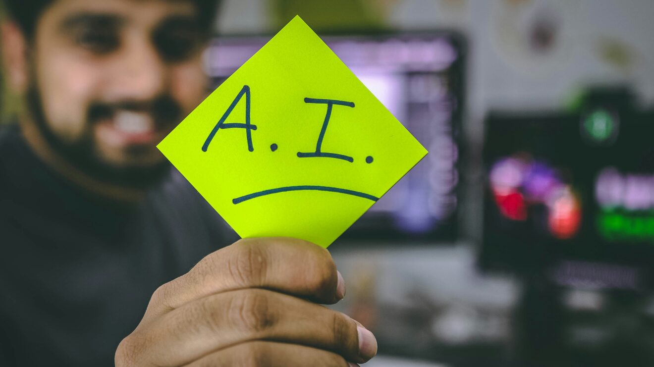 A man behind a computer turning away from it, holding a post-it card with "A.I." on it. Article How generative AI makes scammers alarmingly convincing