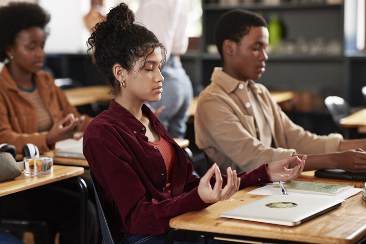 Teenage students with eyes closed meditating at desk in high school classroom for sponsored piece by Calm Classroom