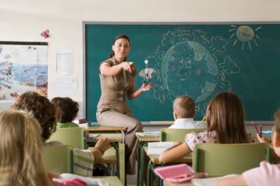 teacher in front of blackboard holding an object and pointing toward students to call on one of them. Thre students at desks are facing her. for education article on coaching tools