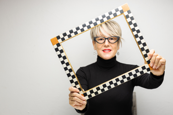 woman with short gray hair in black turtleneck holding empty checkerboard art frame for article on portrait of a staff member