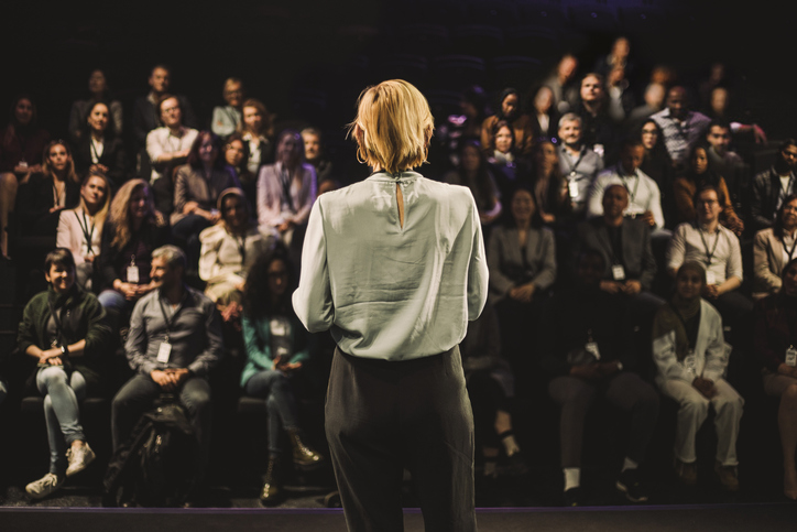 Mature female entrepreneur interacting with audience of professionals wearing lanyards for article on women in education leadership