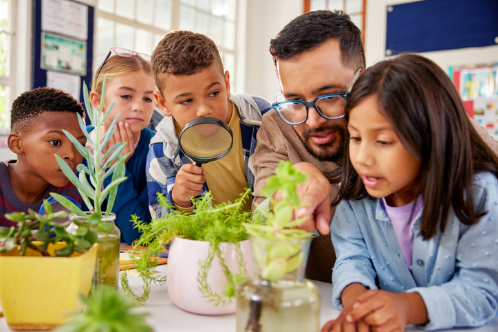 Teacher with schoolchildren in biology class teaching about plants and environment conservation. Little boy holding magnifying glass while observing plant in primary classroom with teacher and classmates. A group of multiethnic school children sitting together and observing a plant with teacher. for sponsored post from Green Ninja on project-based learning