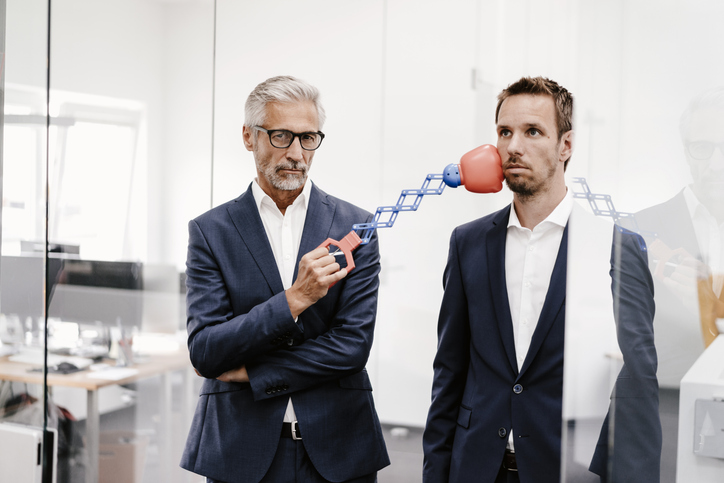 two male workers standing next to each other looking worn out. One has toy accordion-style boxing glove touching the other on the chin. for article on conflict resolution