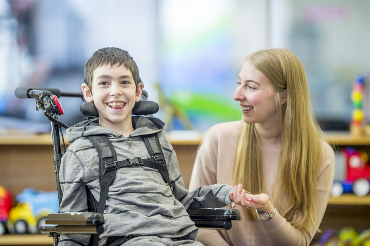 A special needs boy is in his classroom with an assistant. She is holding his hand, and he is smiling at the camera. for education article on IDEA funding